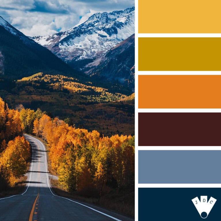 Route sinueuse traversant une forêt aux couleurs d’automne, avec des montagnes enneigées en arrière-plan.